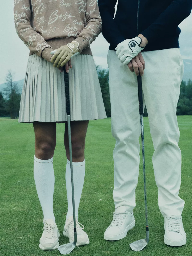 Two models standing on a golf course holding golf clubs, wearing HUGO sportswear including a pleated skirt and white trousers.