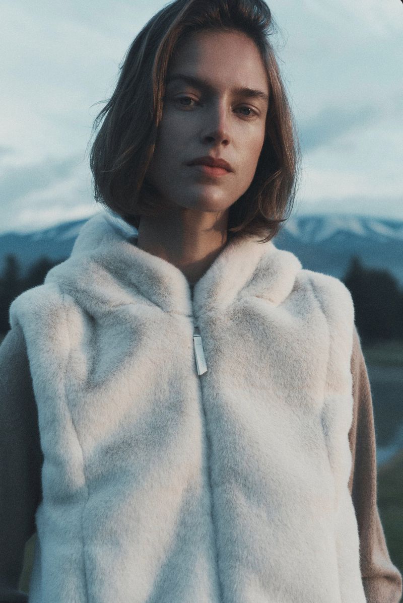 Portrait of a female model wearing a white faux fur vest against an outdoor mountain landscape background under an overcast sky.