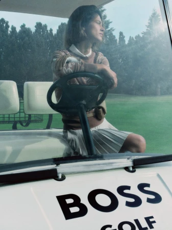 Model seated behind the steering wheel of a golf cart wearing a patterned sweater and pleated skirt for HUGO
