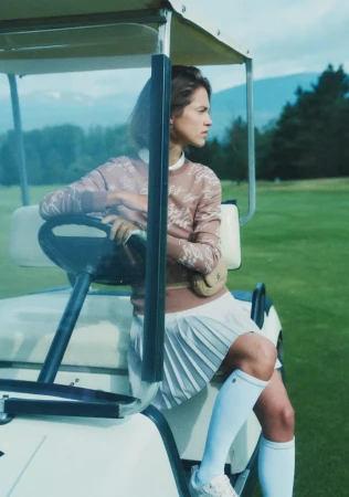 A female model in a golf cart wearing a patterned brown sweater and white pleated skirt on a golf course