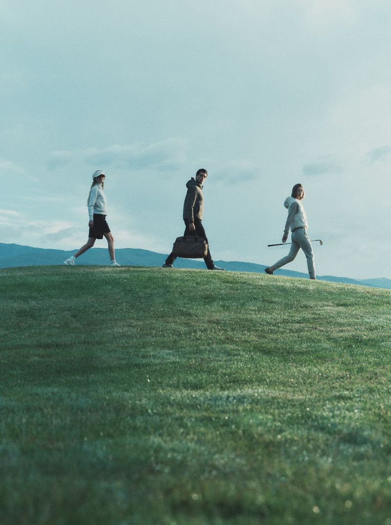Three models walking across a grassy hill carrying a duffel bag and golf club under an overcast sky.