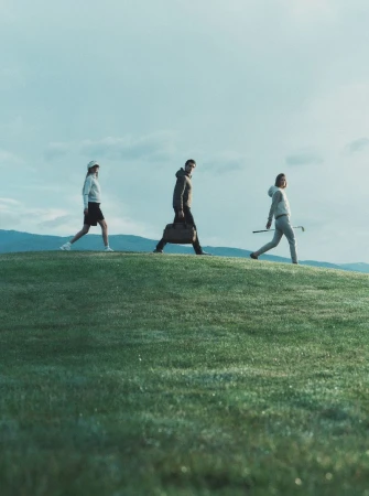 Three models walking across a grassy hill carrying a duffel bag and golf club under an overcast sky.
