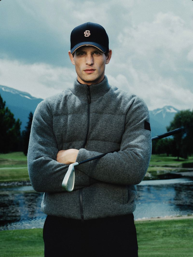 Male golfer wearing a grey puffer jacket and black cap holding a golf club outdoors against a mountain and river background