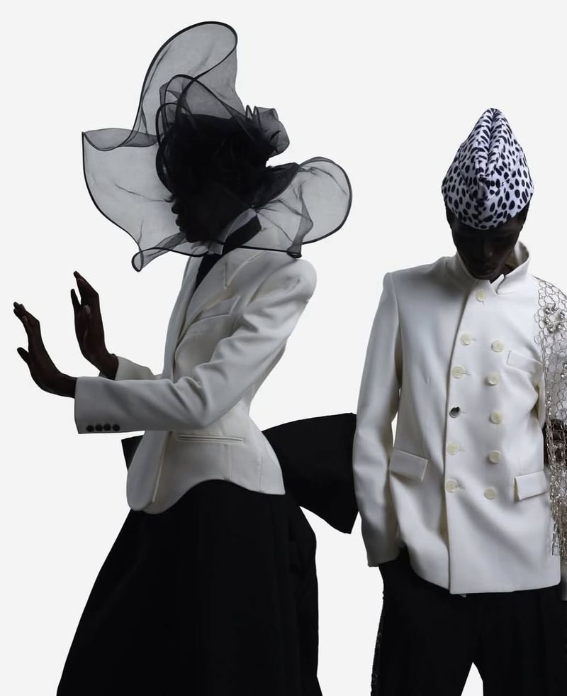 Two fashion models pose in white jackets and black skirts against a white background, one with a large veiled hat, the other with a patterned hat.