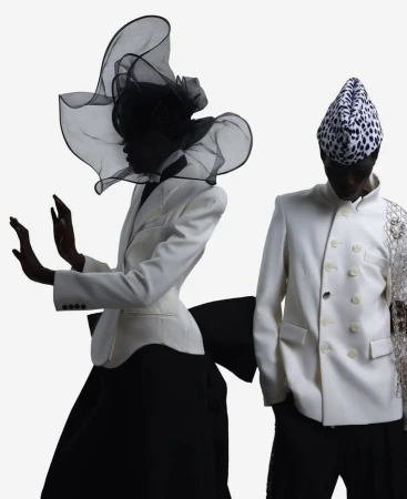 Two fashion models pose in white jackets and black skirts against a white background, one with a large veiled hat, the other with a patterned hat.