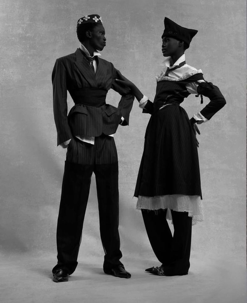 Two models showcase high fashion in a black-and-white studio shot. The model on the left wears a pinstripe suit, while the right model wears a black dress.
