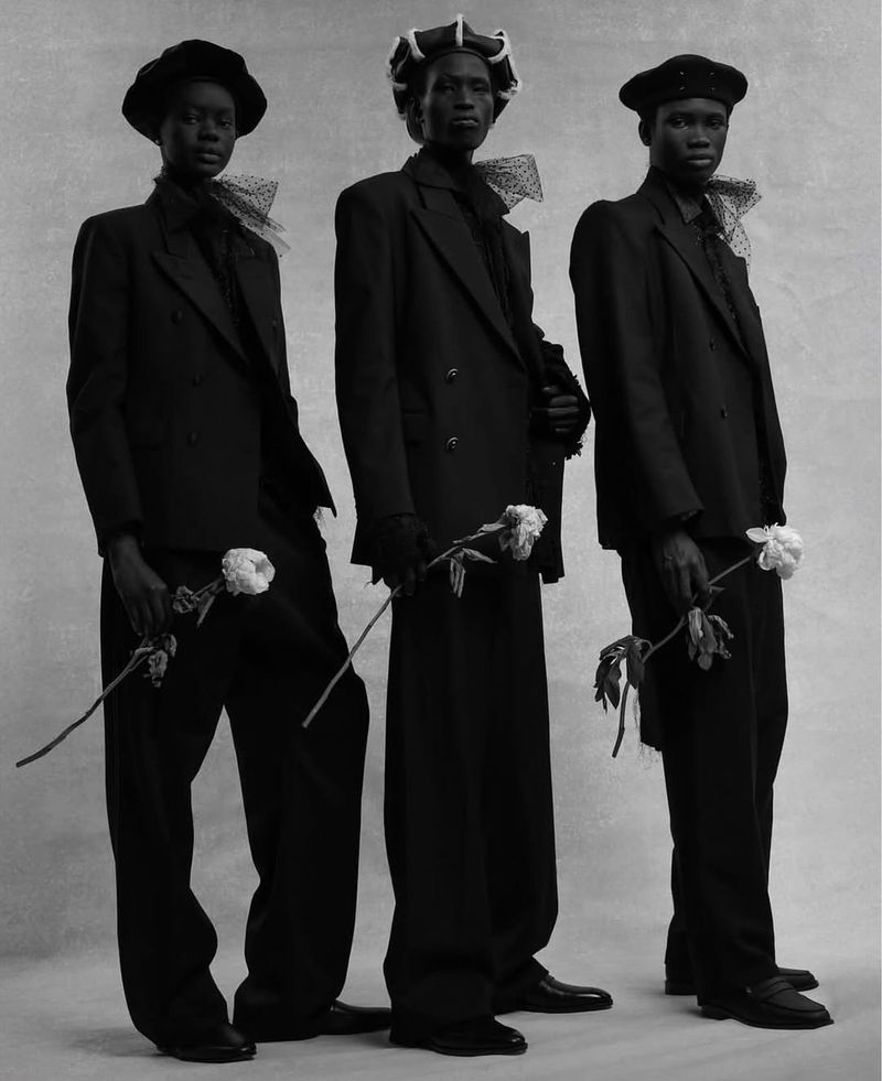 Three Black male fashion models in dark suits and hats hold white roses in a black and white studio portrait with neutral background.
