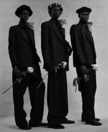 Three Black male fashion models in dark suits and hats hold white roses in a black and white studio portrait with neutral background.