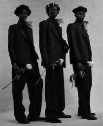 Three models wearing matching black tailored suits and hats holding white flowers against a neutral studio background