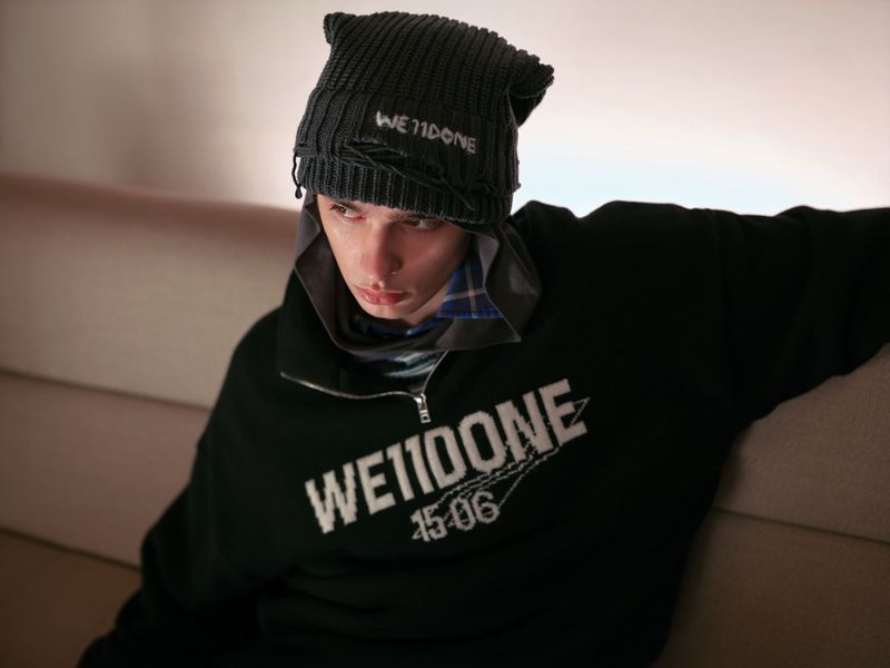 Model wearing a black branded beanie and dark zip-up sweatshirt, seated indoors against a neutral background.