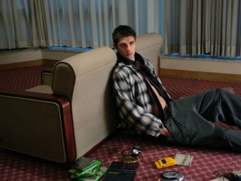 A young male model seated on a patterned carpet leaning against an armchair, wearing a plaid jacket and grey trousers.