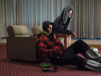 Two models wearing hooded puffer jackets seated in a carpeted interior with patterned curtains and vintage armchairs