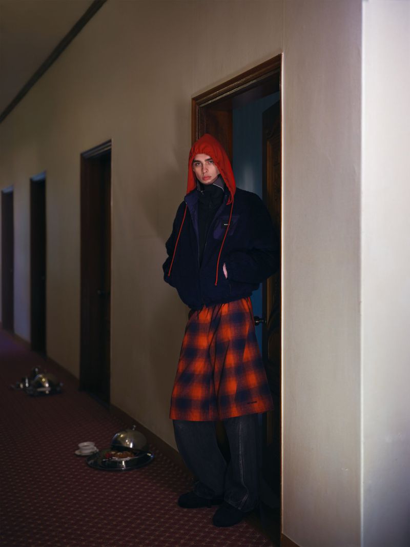 Model standing in a hotel hallway wearing a black jacket with a red hooded layer and plaid skirt for WE11DONE F/W 26 Campaign.