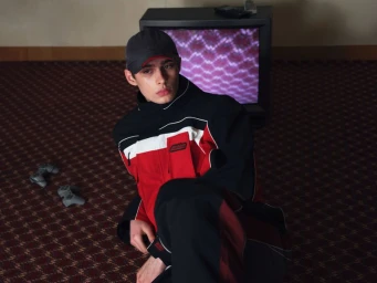 Model seated on a patterned carpet wearing a color-blocked jacket and cap with a vintage television and video game controllers behind him.