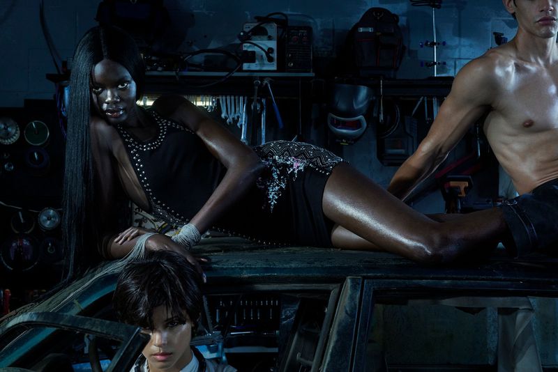 Three models posing in a dimly lit garage setting, featuring a woman lying across the roof of a car.