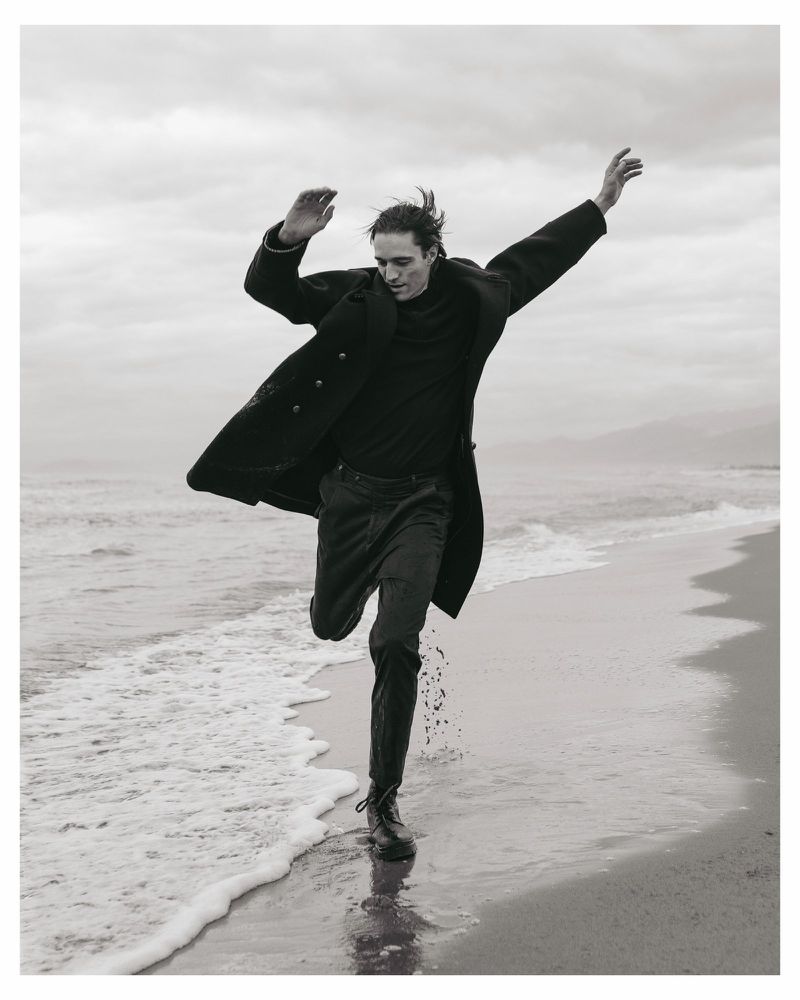 Monochrome image of a man in a long coat leaping along a sandy beach near ocean waves.