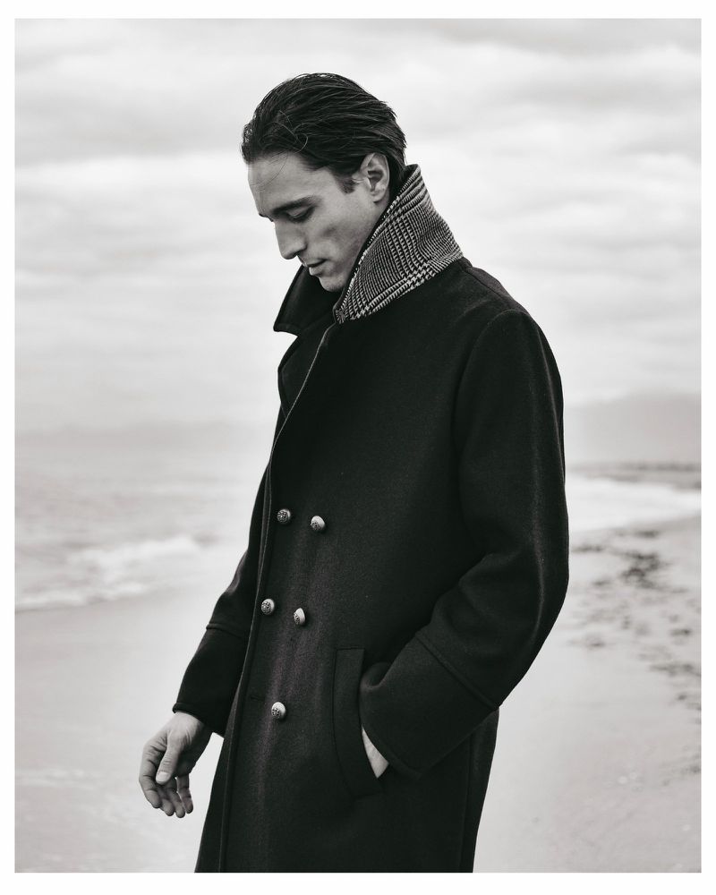 Model Francesco Mariottini in a dark double-breasted coat and patterned scarf on a sandy beach for DISTRETTO12 Autumn 2026