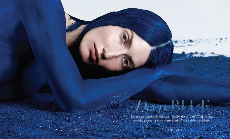 A person lying on a blue textured surface with blue painted skin and dark blue hair, captured in a horizontal editorial portrait.