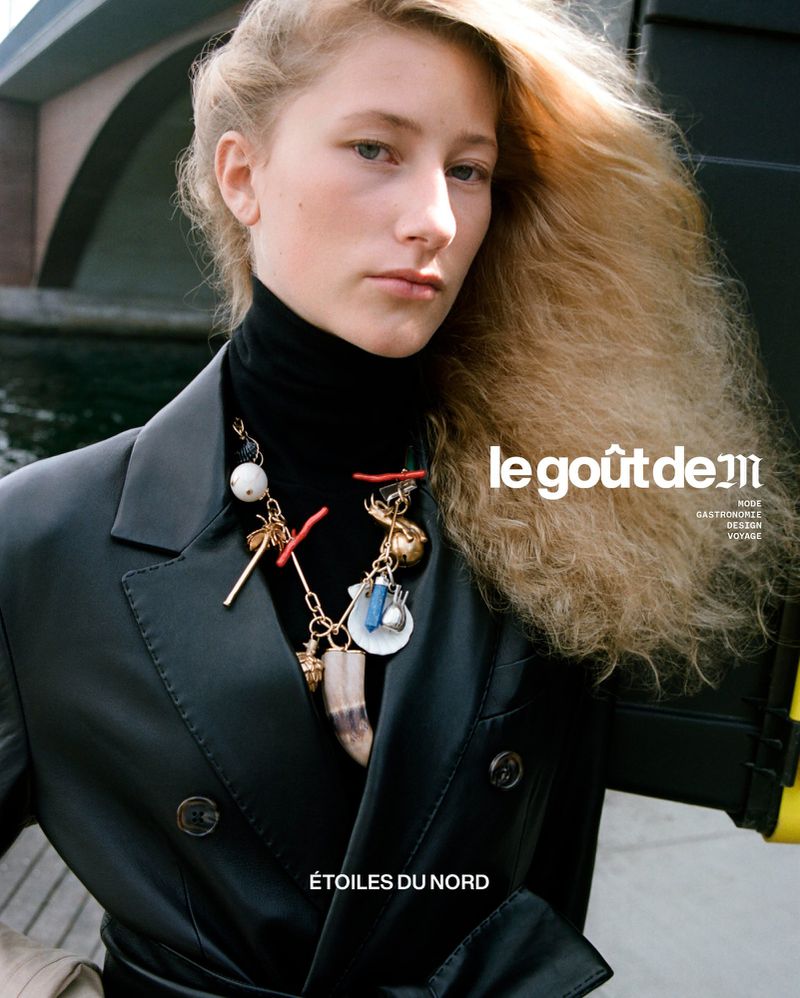 Portrait of a blonde woman wearing a black turtleneck and leather coat with an ornate charm necklace against an outdoor bridge background.