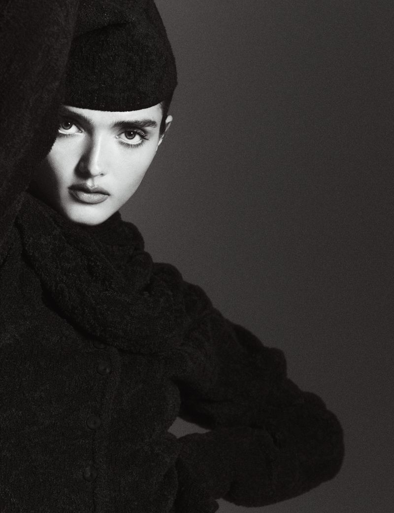 Close-up monochrome portrait of a model wearing a textured dark cap and matching dark wool top against a simple neutral studio background.