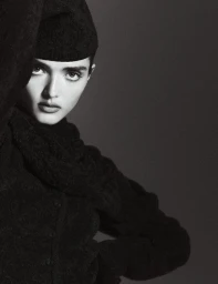 Close-up monochrome portrait of a model wearing a textured dark cap and matching dark wool top against a simple neutral studio background.