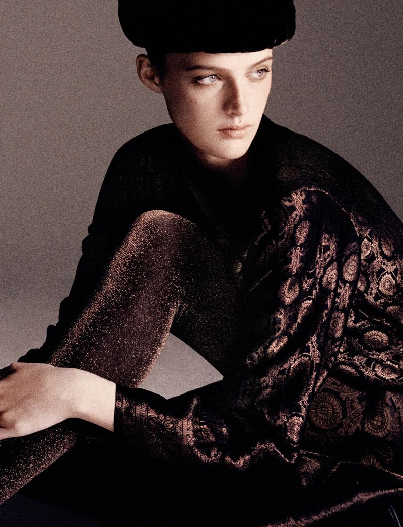 Model wearing a dark patterned top and dark head covering against a neutral grey studio background