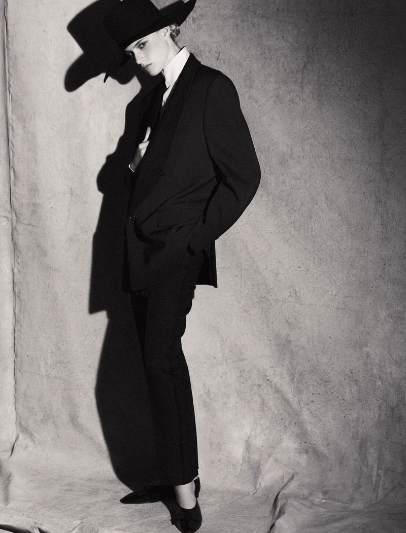 Model in a black suit and wide-brim hat standing against a textured grey backdrop.