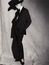 Model in a black suit and wide-brim hat standing against a textured grey backdrop.