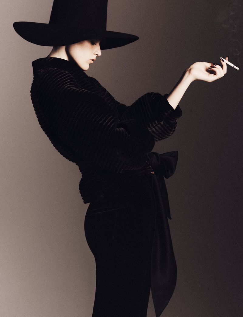 Model in wide-brimmed black hat and dark textured top holding a lit cigarette against a neutral studio backdrop