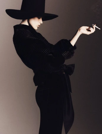 Model in wide-brimmed black hat and dark textured top holding a lit cigarette against a neutral studio backdrop