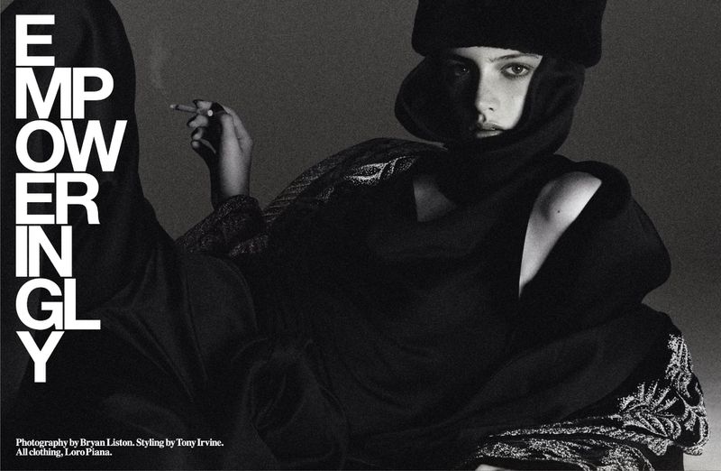 Black and white editorial photograph of a model wearing a dark hooded garment and fur hat, holding a cigarette.