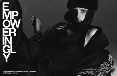 Black and white editorial photograph of a model wearing a dark hooded garment and fur hat, holding a cigarette.