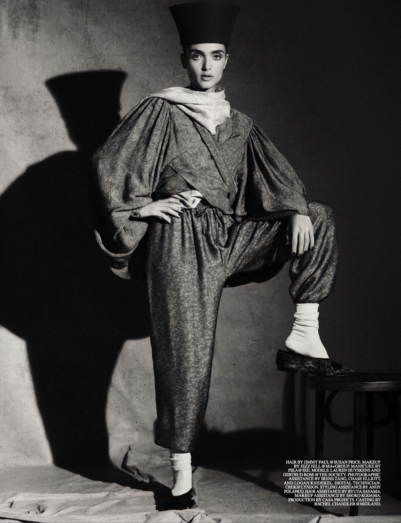 Model in voluminous patterned trousers, matching jacket, tall cylindrical hat and white socks posing on stool