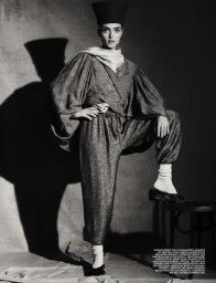 Model in voluminous patterned trousers, matching jacket, tall cylindrical hat and white socks posing on stool