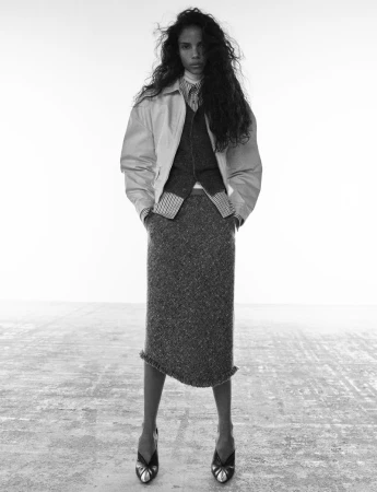 Model with long dark wavy hair wearing a jacket, vest, patterned shirt, and tweed midi skirt on a white studio floor.
