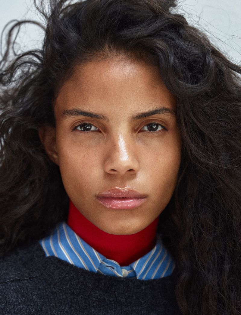 Close-up portrait of a model with dark curly hair wearing a red high-neck collar under a blue striped shirt and dark sweater.