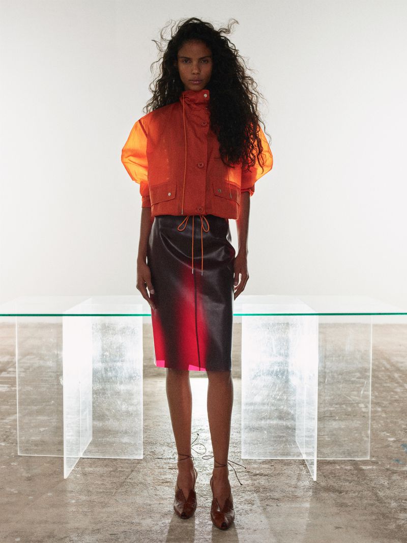 Model standing in an orange jacket and dark skirt before a transparent glass table in a studio setting