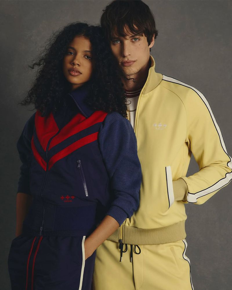 A stylish couple poses in retro tracksuits. The woman wears a navy and red tracksuit, while the man sports a yellow tracksuit.
