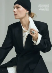 Model wearing a black blazer, white shirt, and black beanie holding her collar against a grey background