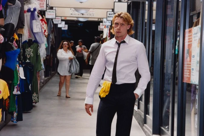 George Mackay walking through an indoor arcade wearing a white shirt and dark pinstripe trousers