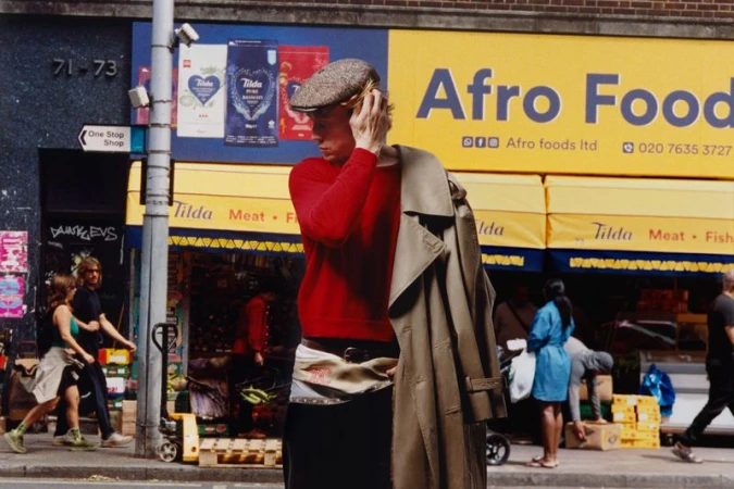George MacKay wearing red sweater and flat cap holding trench coat outside Afro Foods shop