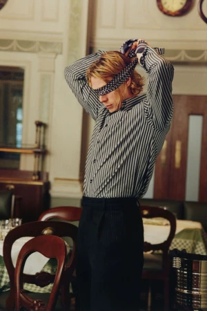 George Mackay tying a patterned blindfold over his eyes, wearing a striped shirt and dark trousers inside a restaurant.