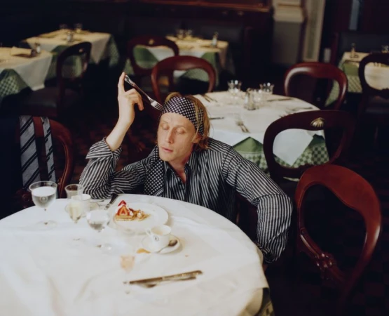 George Mackay wearing a patterned headband and striped shirt seated at a table in a restaurant for GQ Magazine