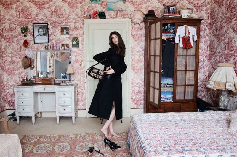 Woman in a black coat holding a patterned handbag in a bedroom with red toile wallpaper, standing barefoot on a patterned rug.