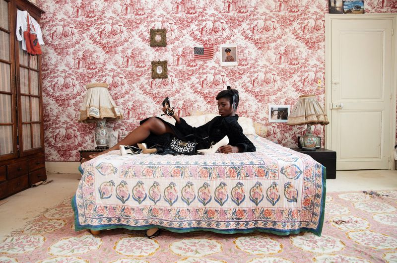 Black woman lounging on a bed in a patterned room holding a shoe for AnOther Magazine editorial