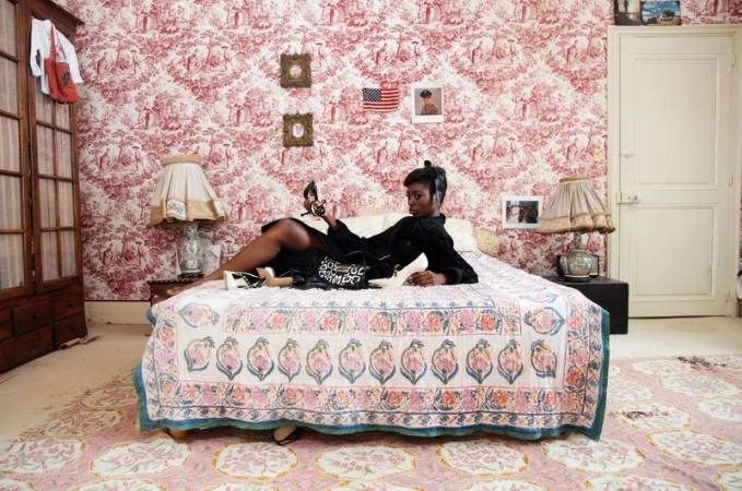 Black woman lounging on a bed in a patterned room holding a shoe for AnOther Magazine editorial