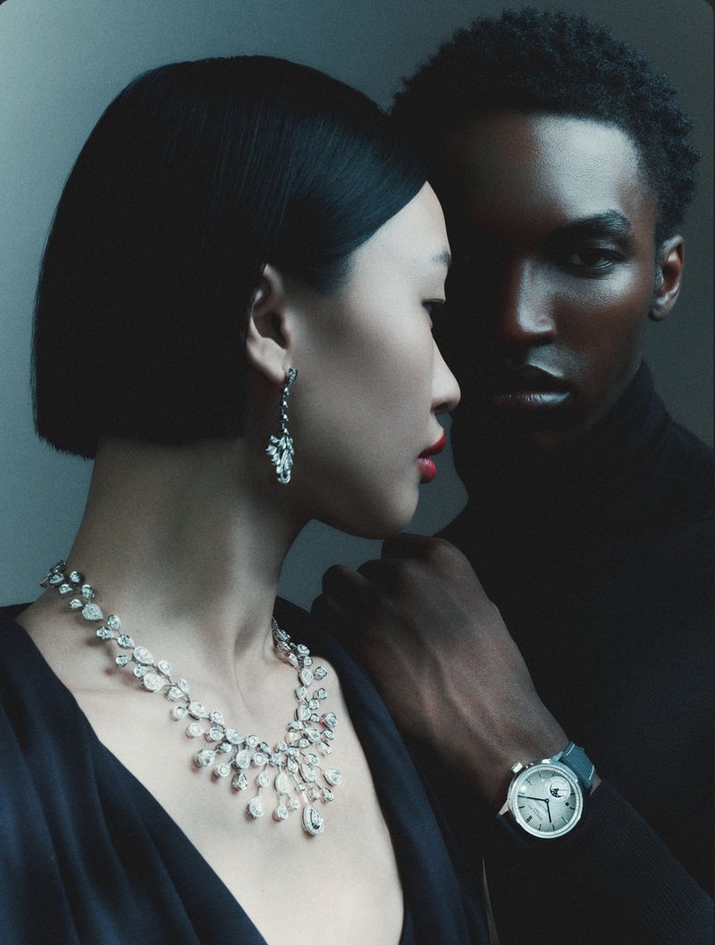 Close-up portrait of two models wearing fine jewelry and a watch against a dark grey seamless studio background.