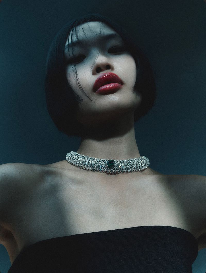 Close-up portrait of a woman wearing a heavy jeweled choker necklace and a dark strapless top against a moody background.