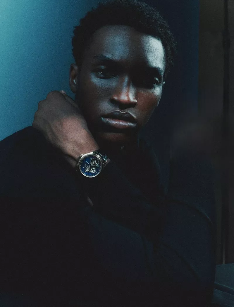 Close-up portrait of a Black man wearing a black shirt and a luxury watch against a dark blue background