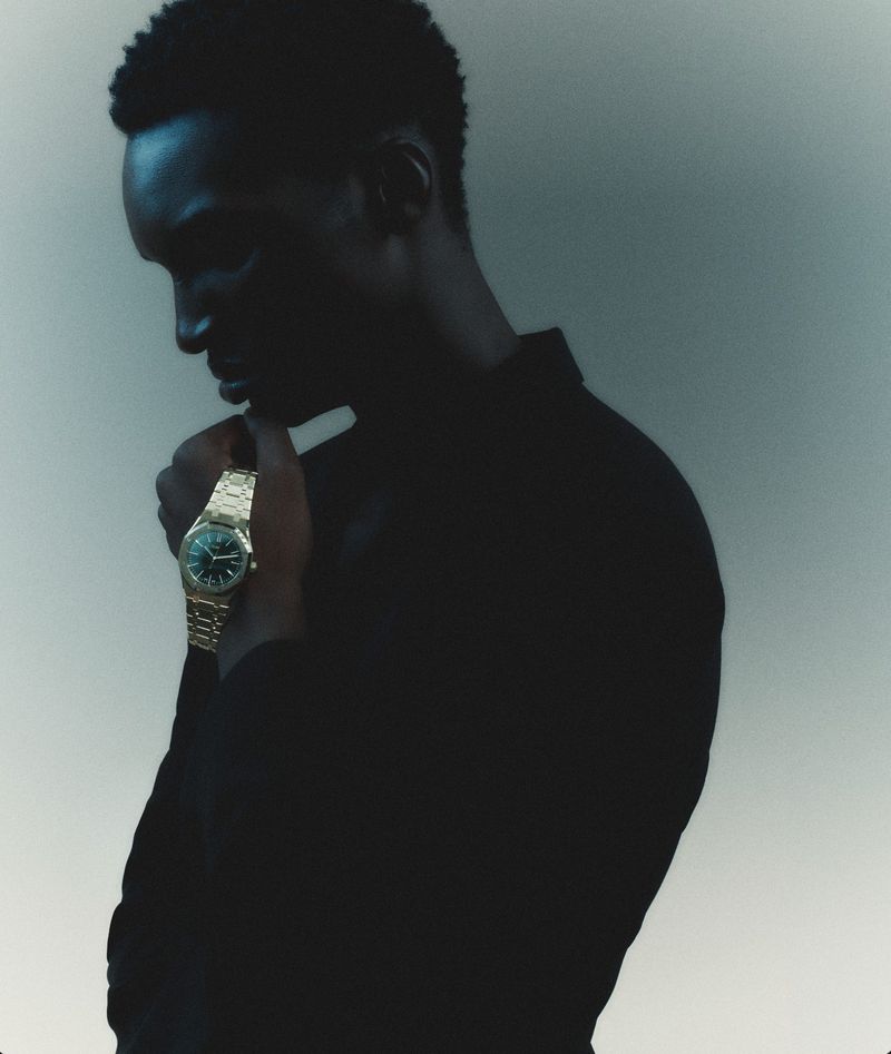 Black man wearing a gold watch and dark shirt posing against a plain grey background